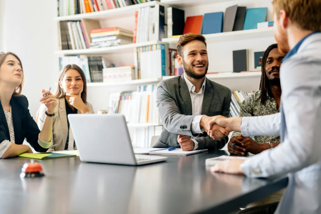 Team meeting in a bright office; two colleagues shake hands as others smile, with bookshelves in the background.